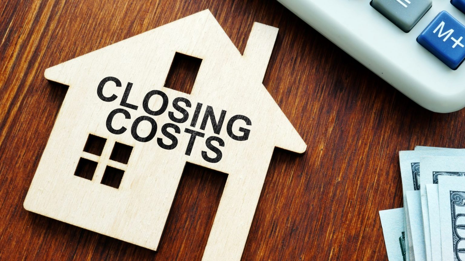 Wooden house laying on brown wooden table that reads "CLOSING COSTS"