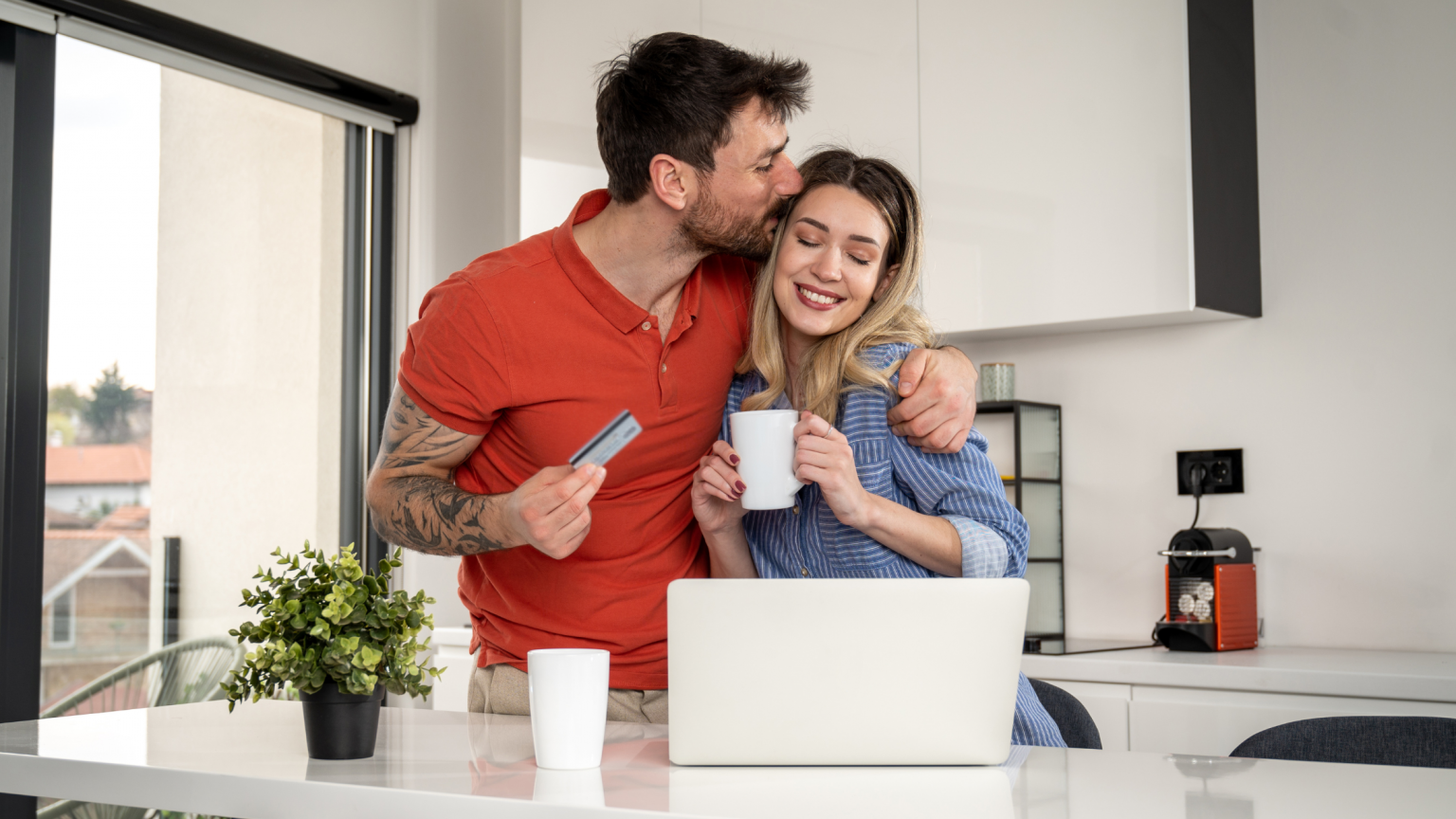 Happy young couple in kitchen. Young male kissing side of head of young female, holding cup of coffee in kitchen with laptop on counter.