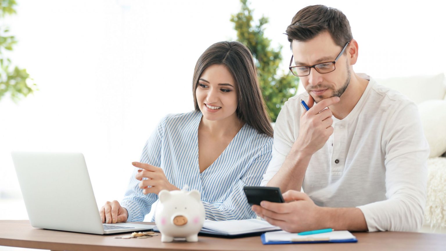 Young male and female couple looking at laptop and discussing money at a table. Male holding calculator. Piggy bank on table.