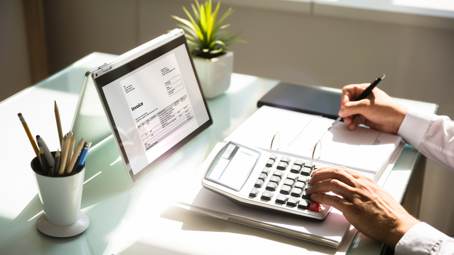 Woman using calculator to determine home affordability, working on desk with laptop and plants and pencils.