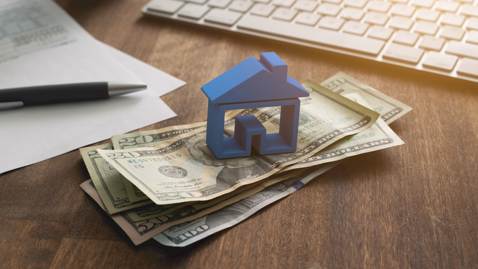 Blue house figurine sitting on a stack of $20 dollar bills on a desk.